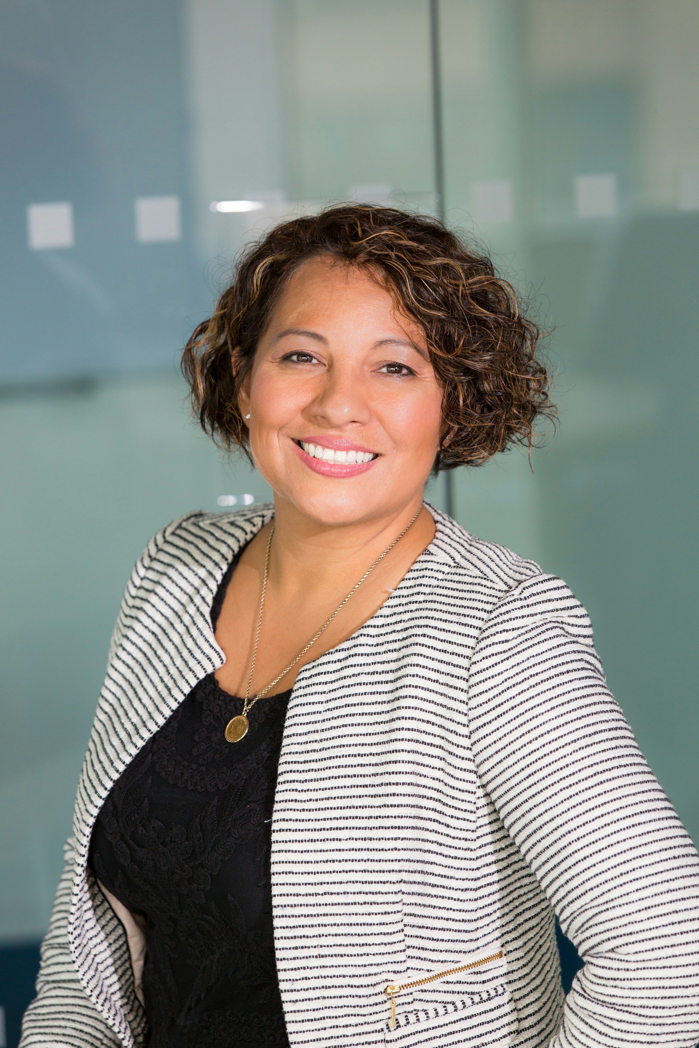 Lächelnde Frau mit Locken und gestreifter Strickjacke vor einer Glaswand im Büro